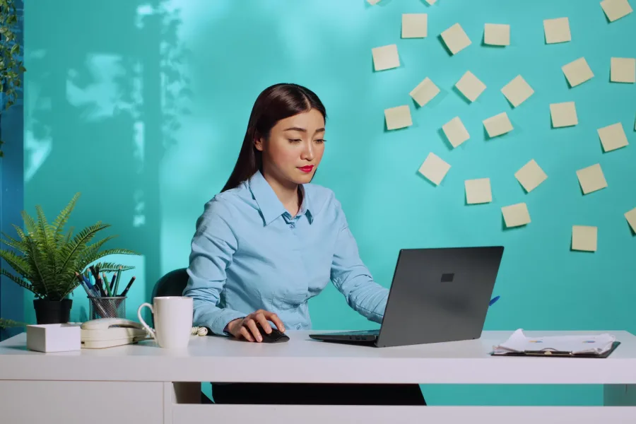 Professional businesswoman bored in administration office, playing with pen while working on laptop. Fatigued employee woman working in modern workplace over blue studio background